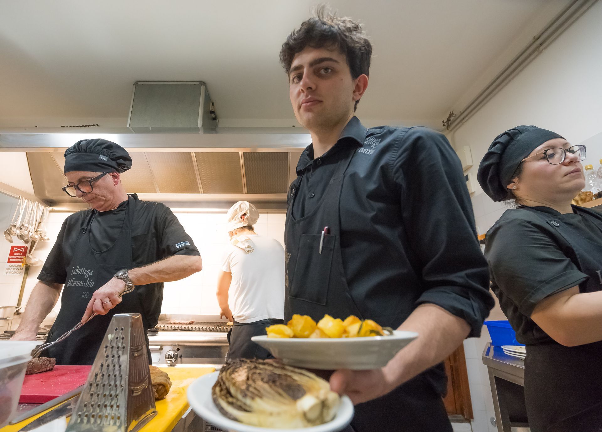 Cuochi in cucina che preparano il cibo; uno tiene in mano un piatto di patate, un altro affetta la carne.