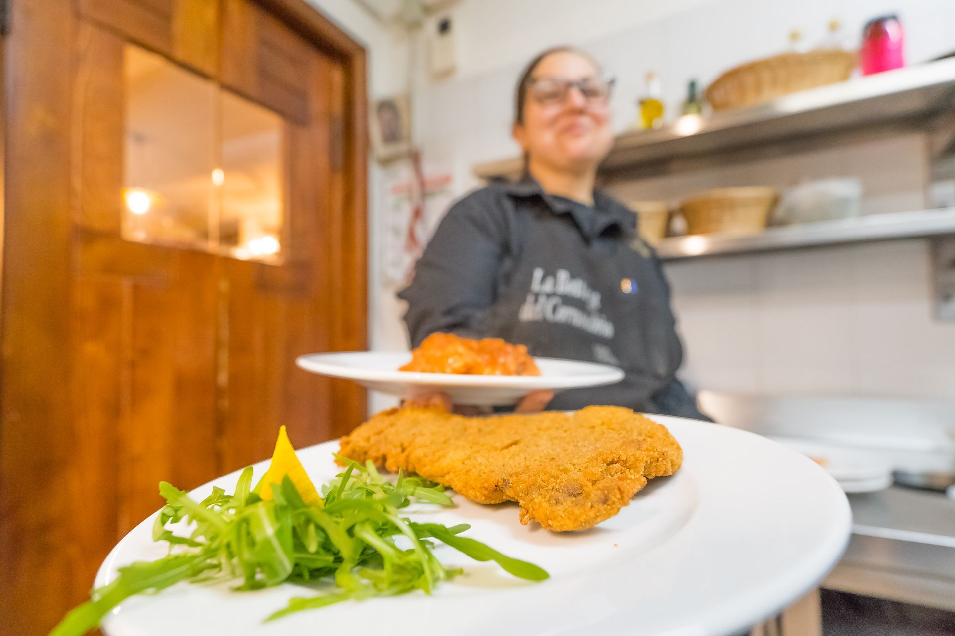 Donna che tiene in mano due piatti di cibo, uno con carne impanata e verdure, l'altro con una salsa, nella cucina di un ristorante.