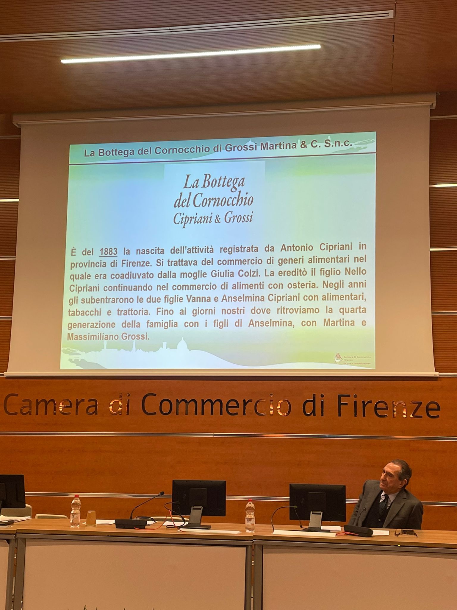 Un uomo seduto a un tavolo da conferenza con uno schermo che mostra il testo: 