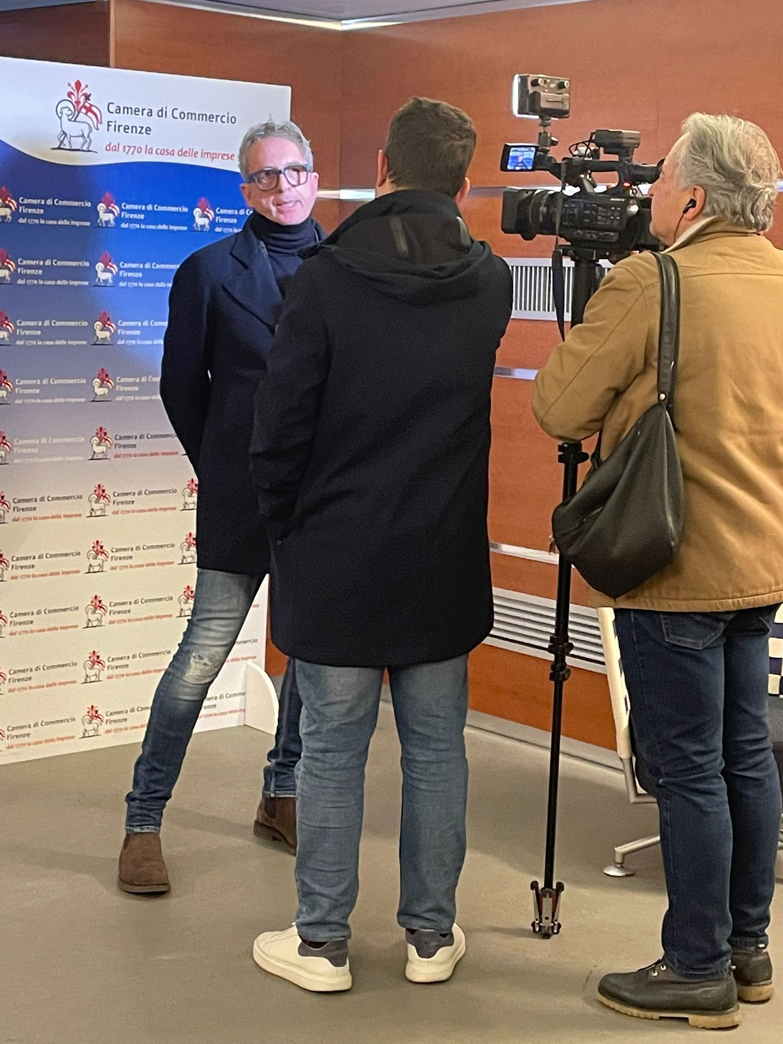 Un uomo con gli occhiali viene intervistato da una persona con una telecamera, mentre un'altra persona è in piedi nelle vicinanze. All'interno.