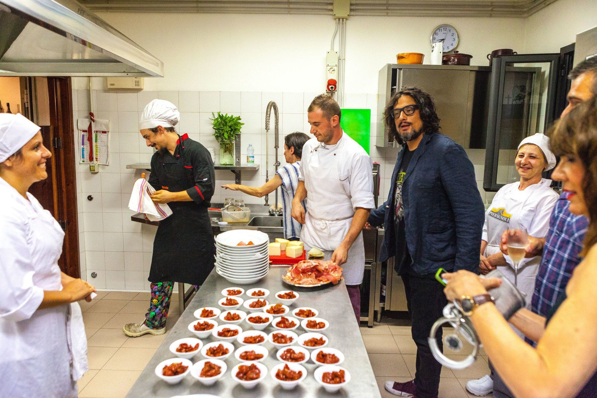 Chef e ospiti in cucina osservano il cibo preparato. Piano di lavoro in acciaio inox con ciotole.
