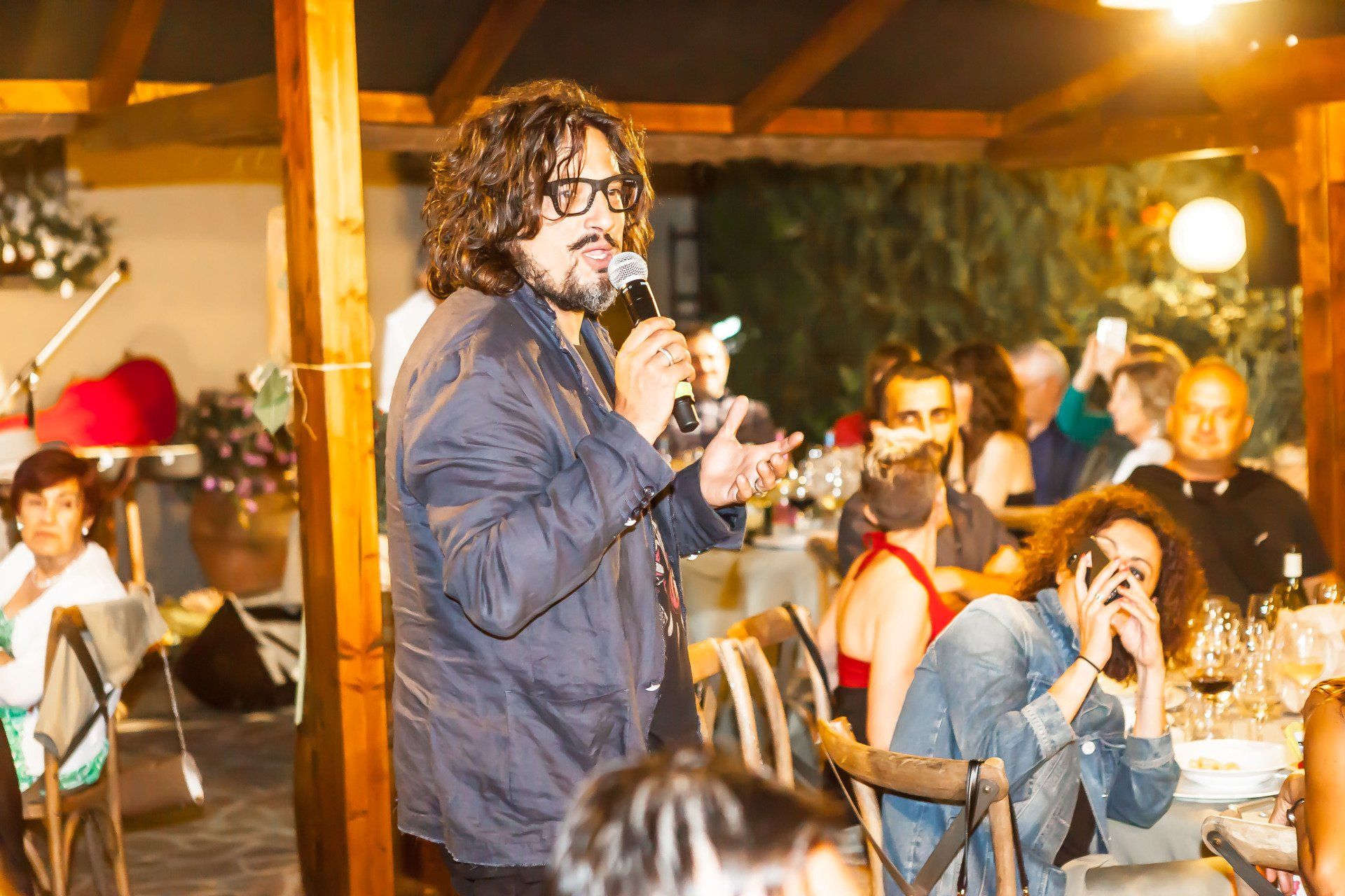 Un uomo con i capelli ricci e gli occhiali parla al microfono durante un evento all'aperto. Gli ospiti sono seduti ai tavoli.
