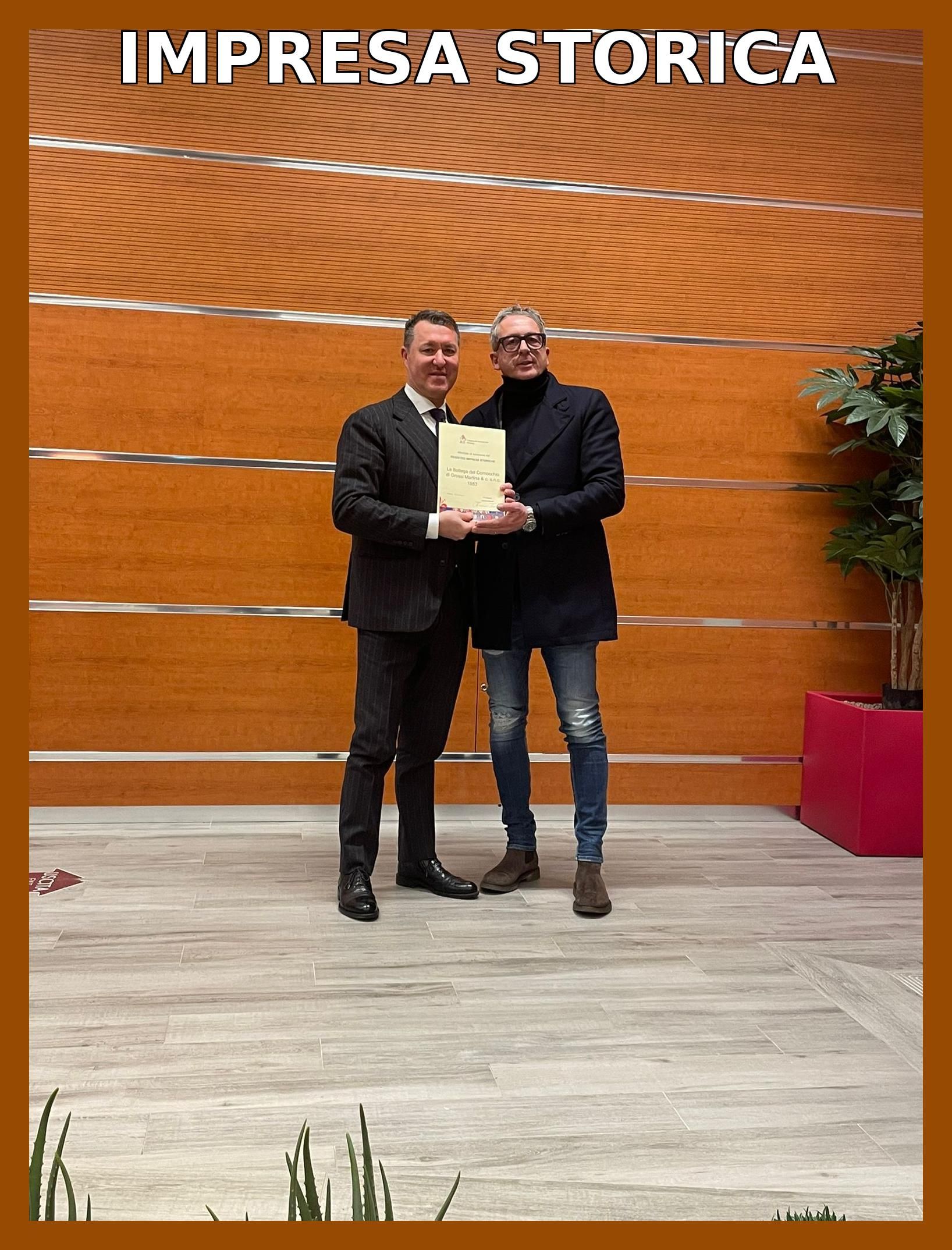 Due uomini tengono in mano un documento, in un edificio con pannelli di legno. In alto la scritta 