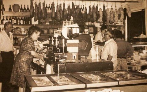 All'interno di un negozio, persone sedute al bancone con una vetrina, cibo appeso in alto e bottiglie sugli scaffali.