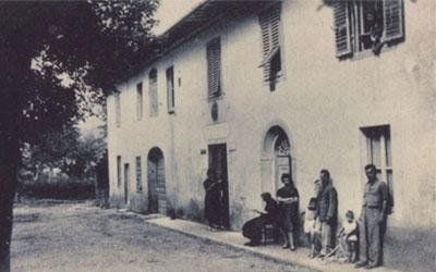 Foto in bianco e nero di un edificio a due piani con diverse persone in piedi all'esterno, probabilmente in un villaggio europeo.