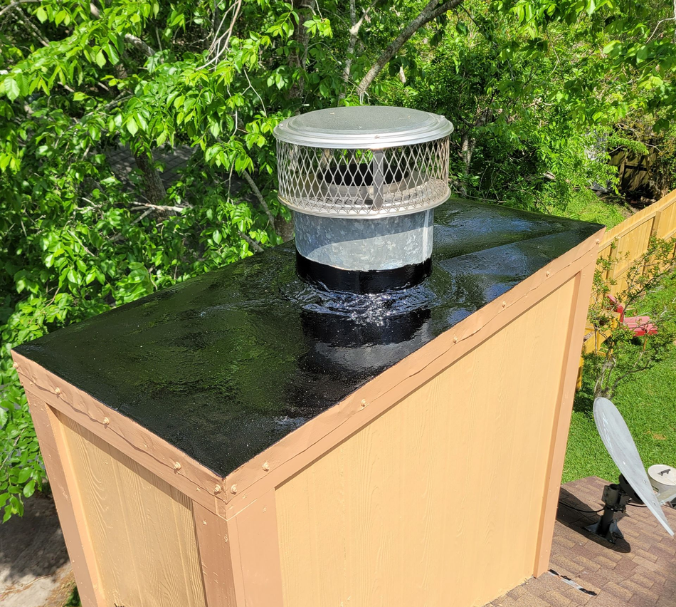 Outdoor rooftop vent chimney on a tan utility box with a black top, surrounded by green trees