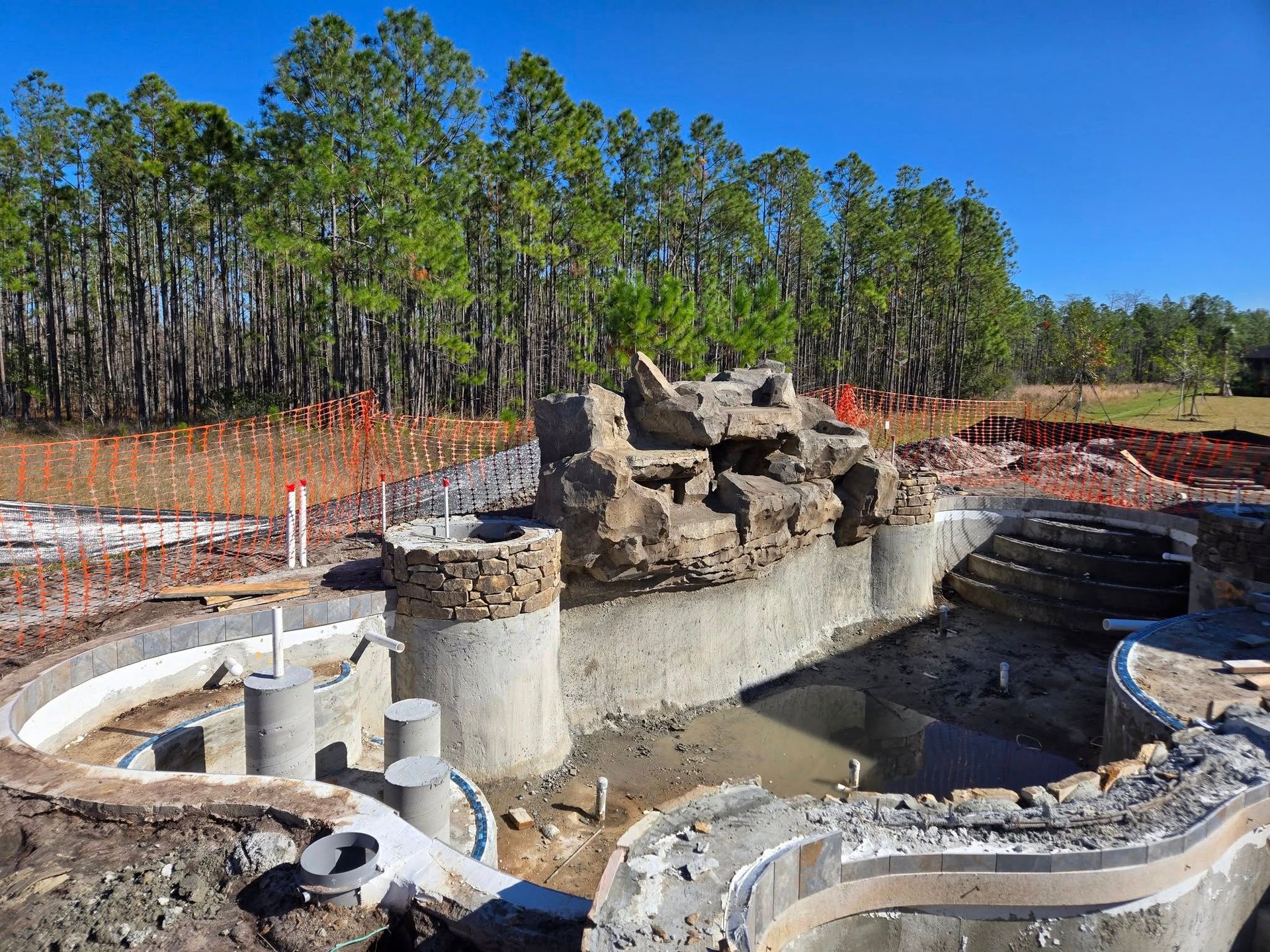 Pool construction site with concrete structures, rock feature, and trees in background.