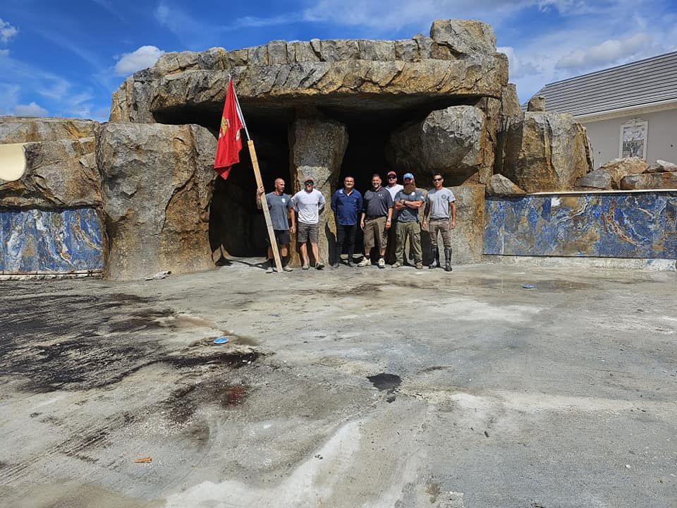 Group of men standing in front of a rock structure with a flag, outdoors.
