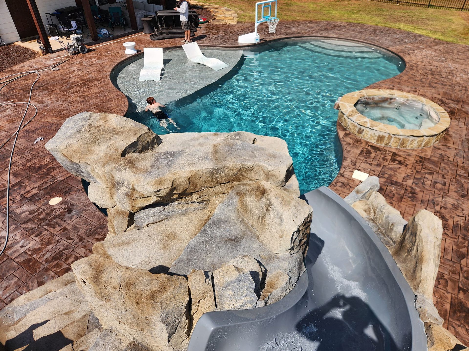 A swimming pool with a rock slide, hot tub, and two lounge chairs on a stamped concrete deck.