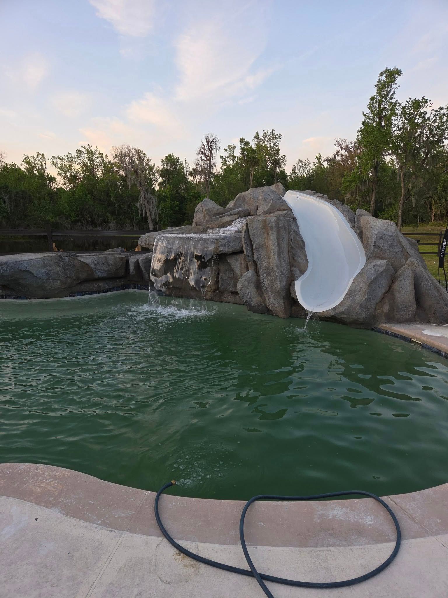 A pool with a rock waterfall and water slide. The water is green. Trees in the background.