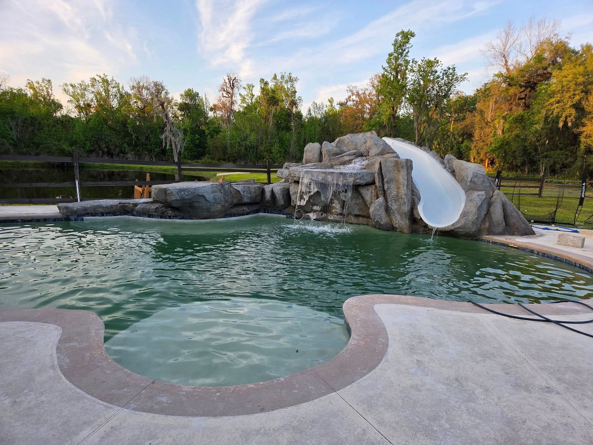 Swimming pool with water slide, rock waterfall feature, and surrounding trees under a blue sky.