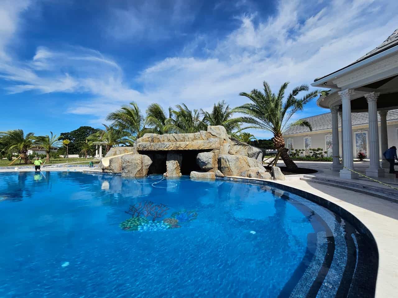 Pool with rock waterfall, palm trees, and ornate building under a bright blue sky.