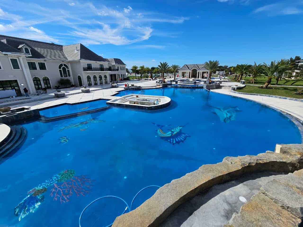 Luxurious estate with large blue pool; a mansion and palm trees under a bright blue sky.