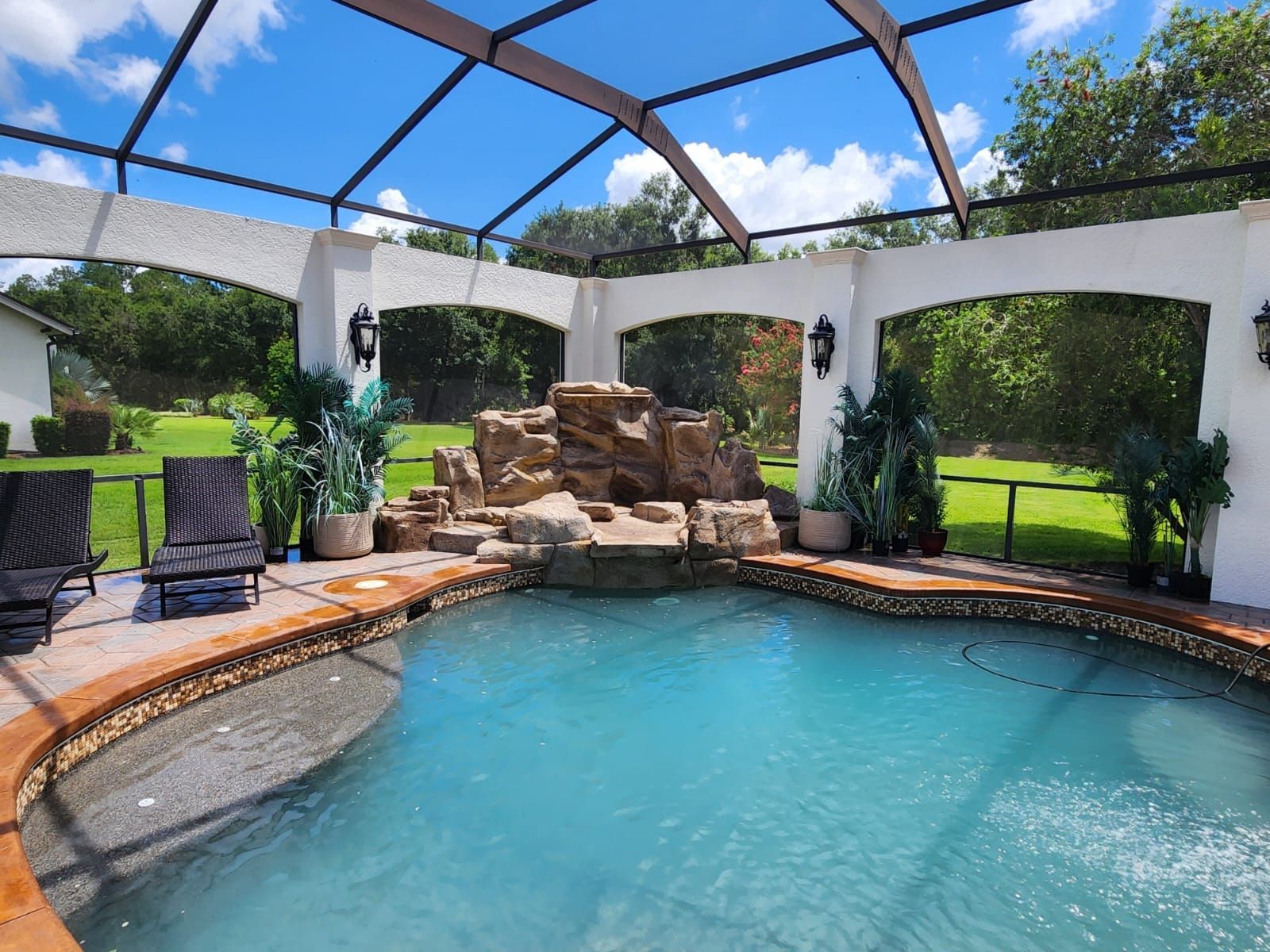 Swimming pool with rock waterfall feature, covered patio, lush green lawn, sunny day.
