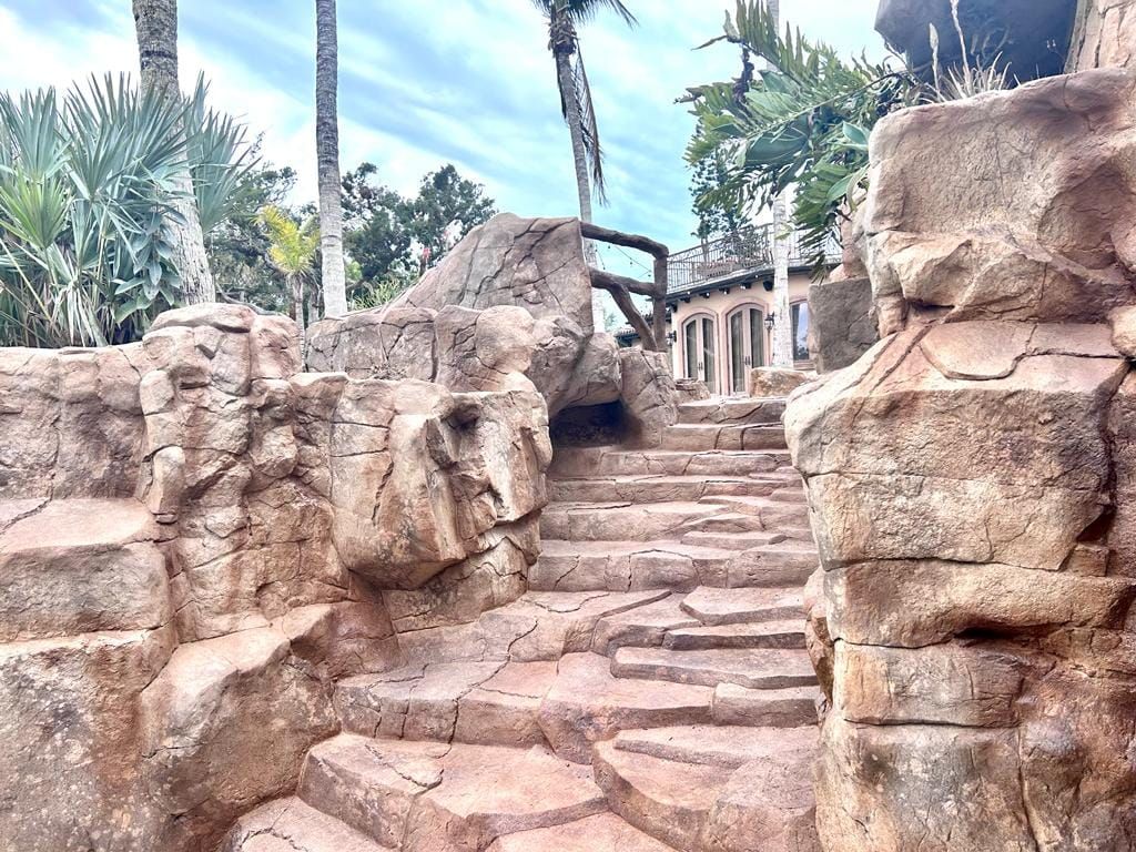 Stone steps leading up, framed by large rocks, to a building with columns and palm trees.