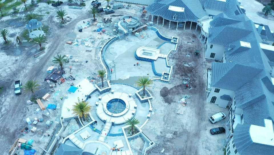 Aerial view of a luxury pool area under construction with multiple pools, structures, and palm trees.