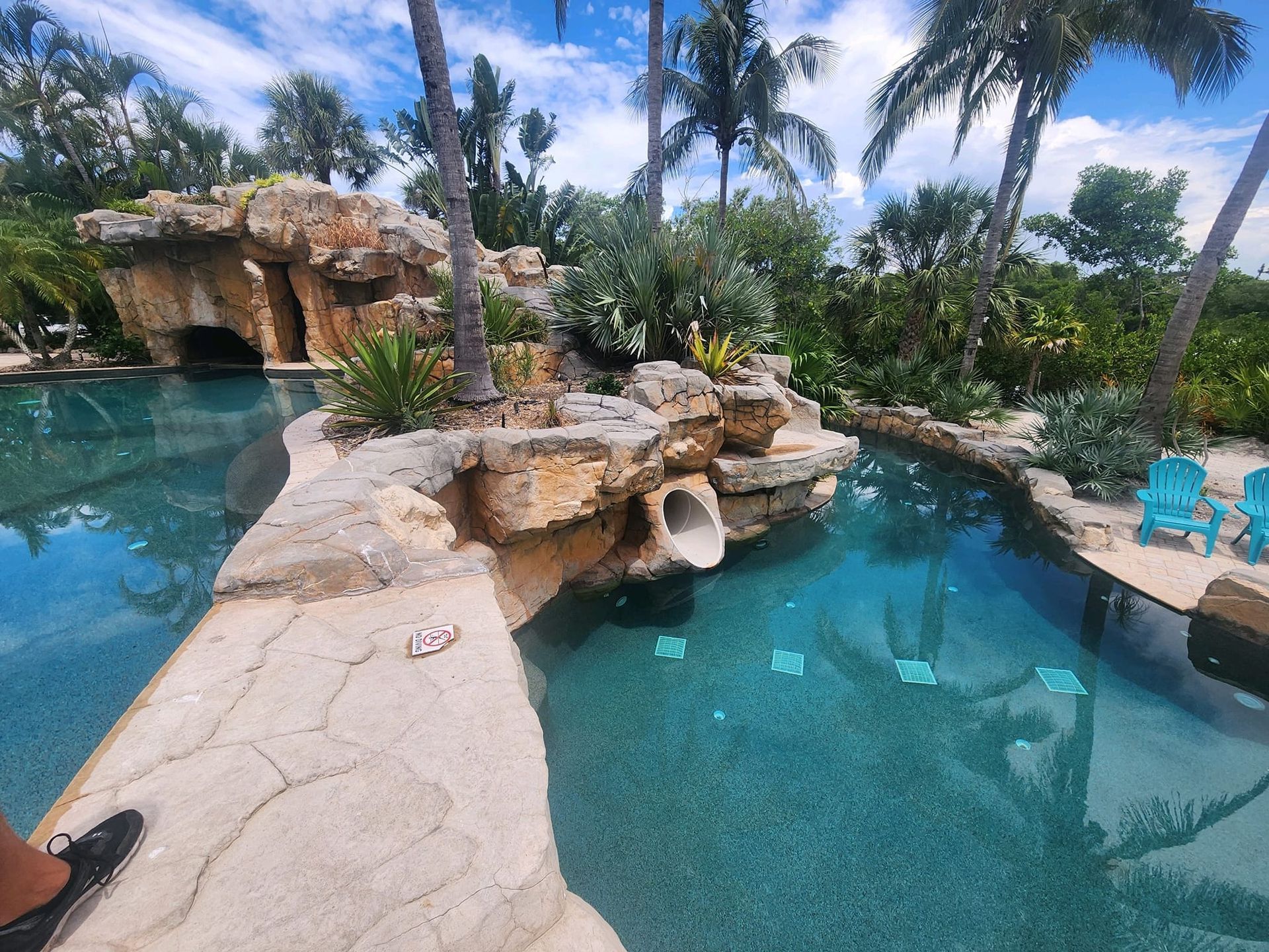 Pool with rock formations, waterfall, palm trees, and blue chairs. Clear blue water and sunny sky.
