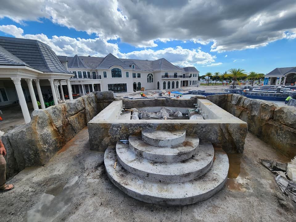 Construction of a stone-lined pool with circular steps. A large house is in the background under a cloudy sky.