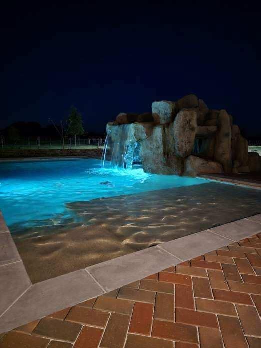 Nighttime pool with illuminated blue water and rock waterfall feature.