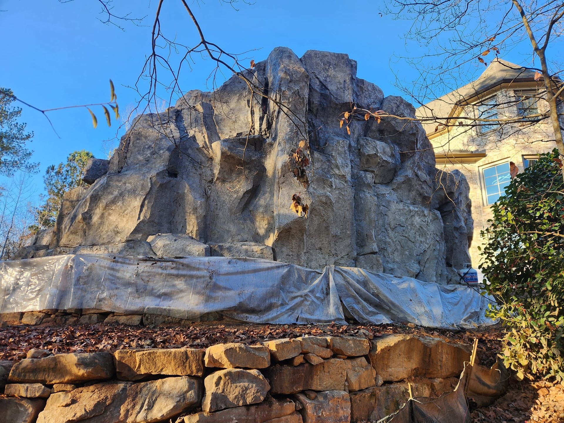 Artificial rock structure on a stone wall, in front of a house. Tarpaulin at its base. Sunny day.