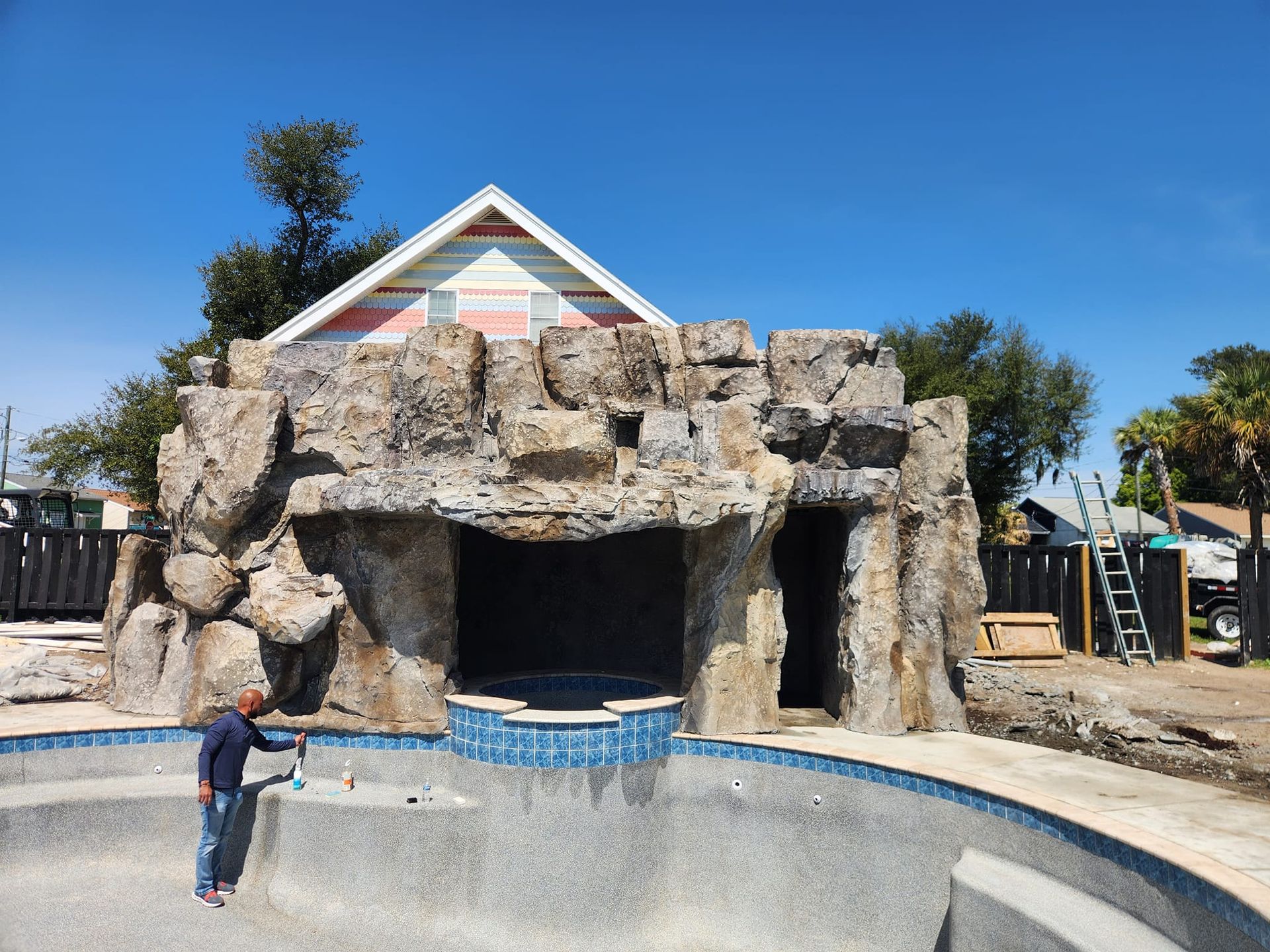 Pool with rock waterfall feature, man working on pool. House with peaked roof in the background. Blue sky.