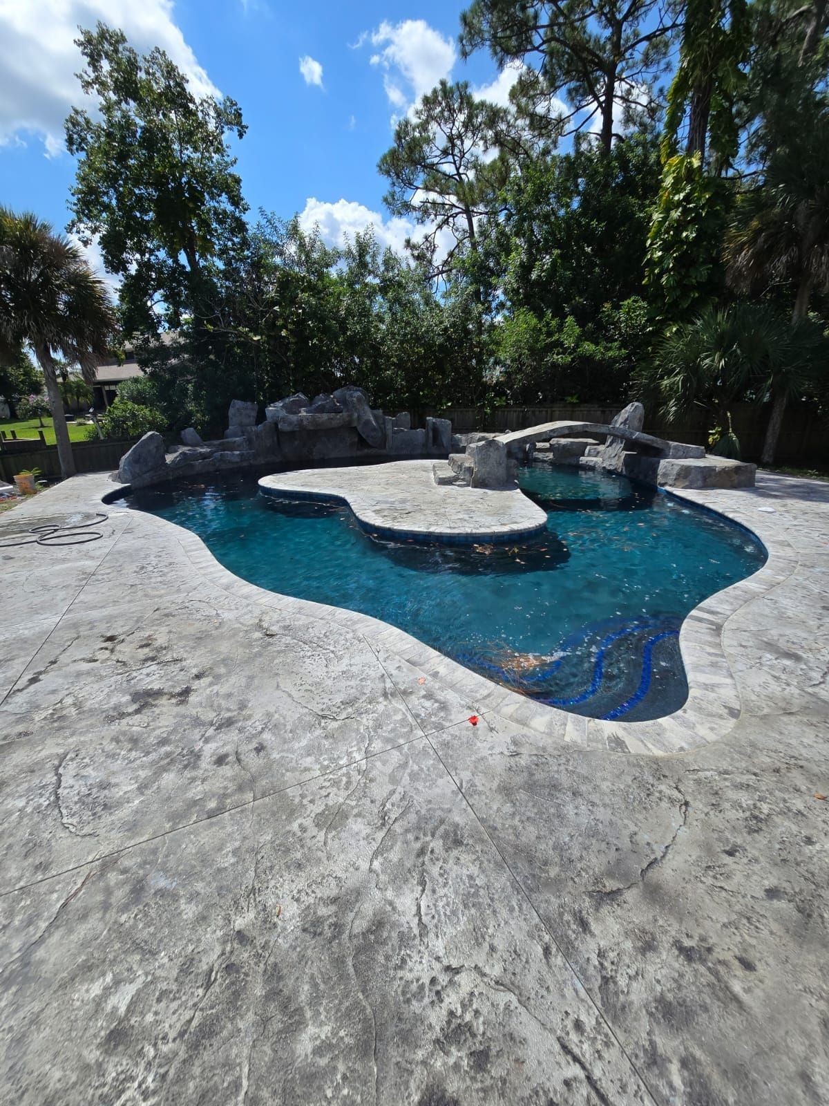 A freeform swimming pool with a rock waterfall surrounded by textured concrete, with trees in the background under a blue sky.