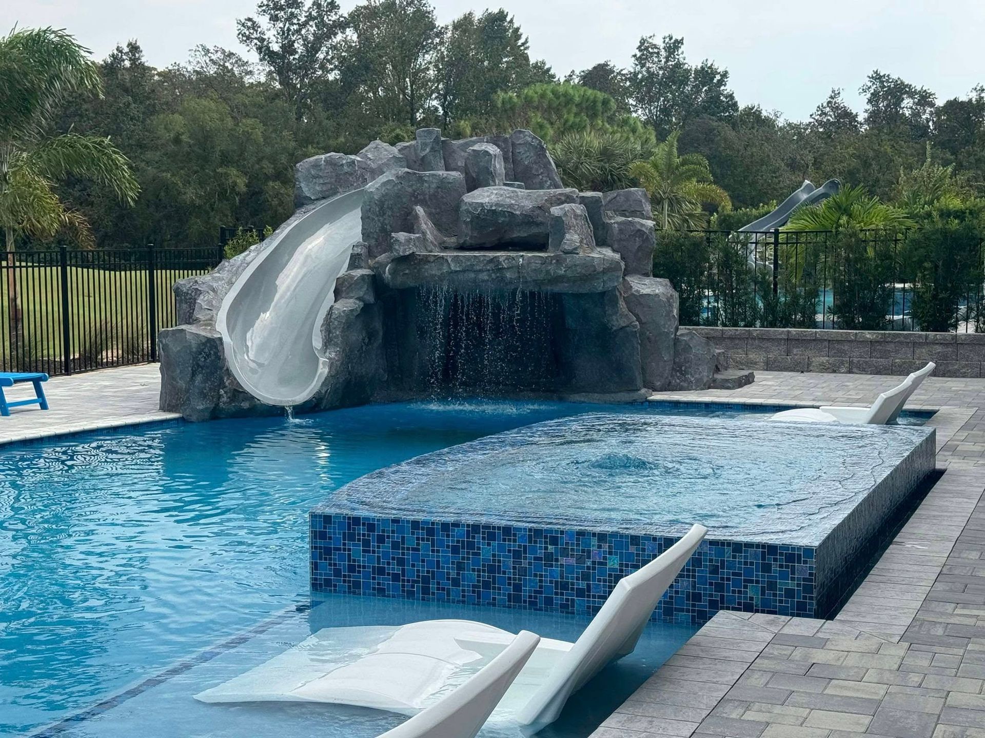 Swimming pool with a water slide and waterfall feature. Blue water, stone accents, and surrounding trees.