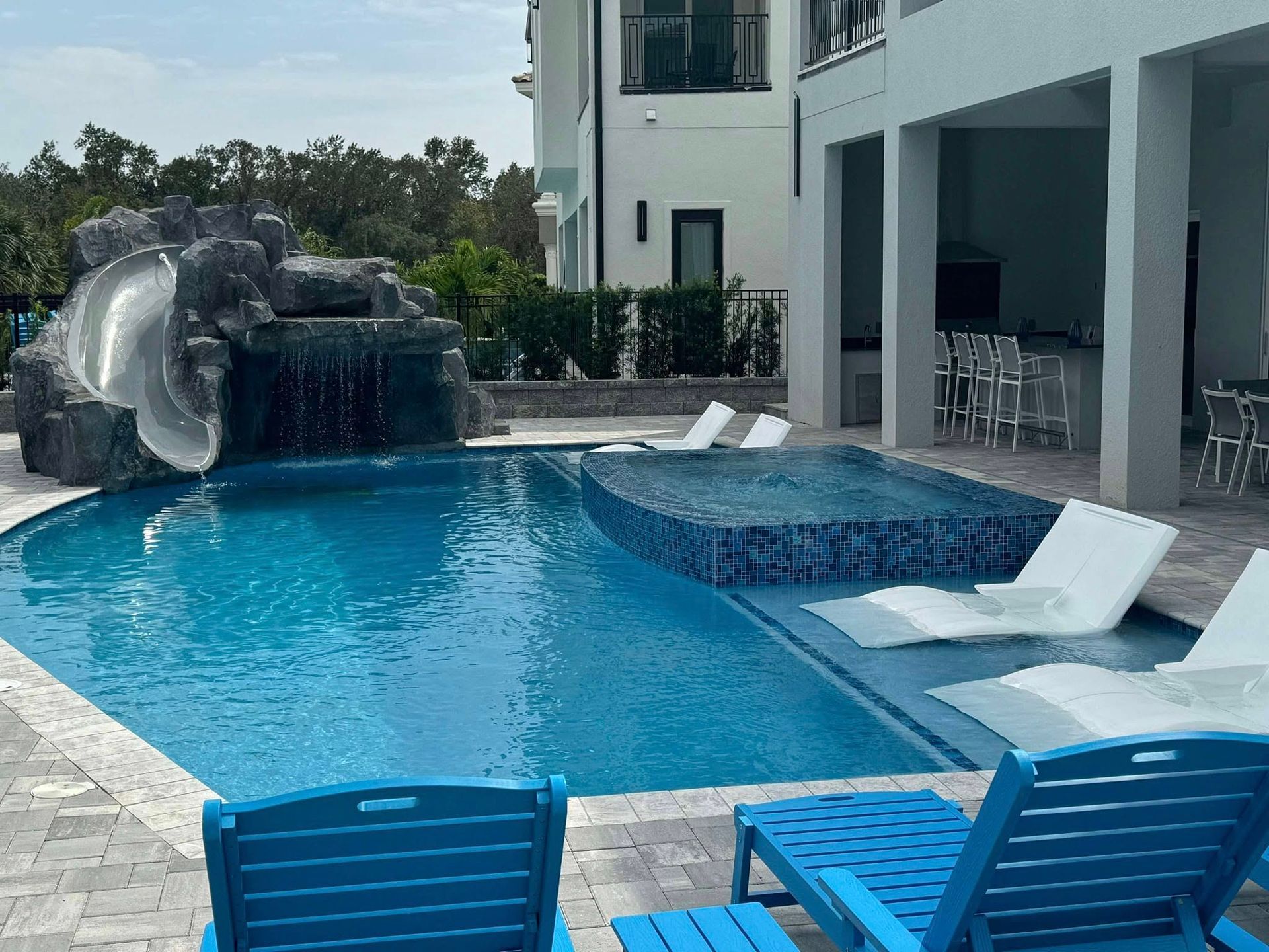 Swimming pool with waterfall slide, hot tub, lounge chairs, and a modern home.