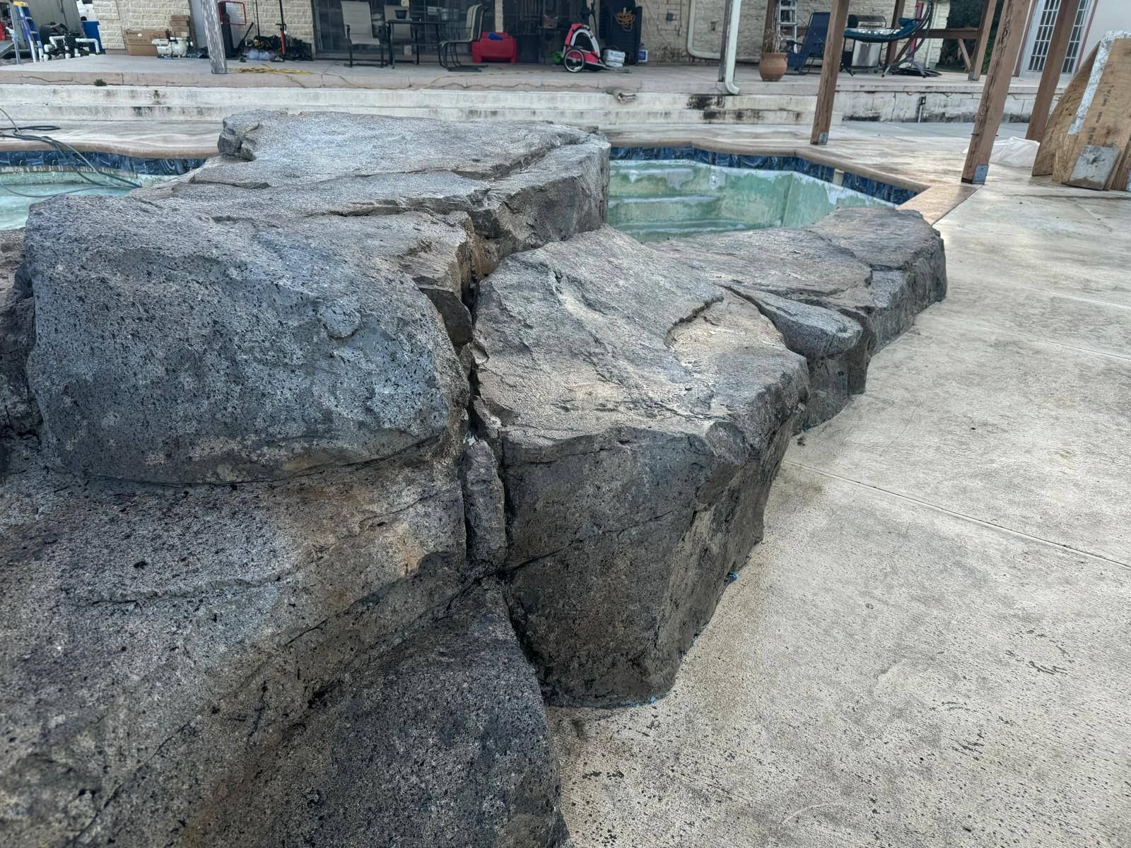 Large, dark rock formation beside a pool. Concrete patio in foreground; light-colored pool water visible.