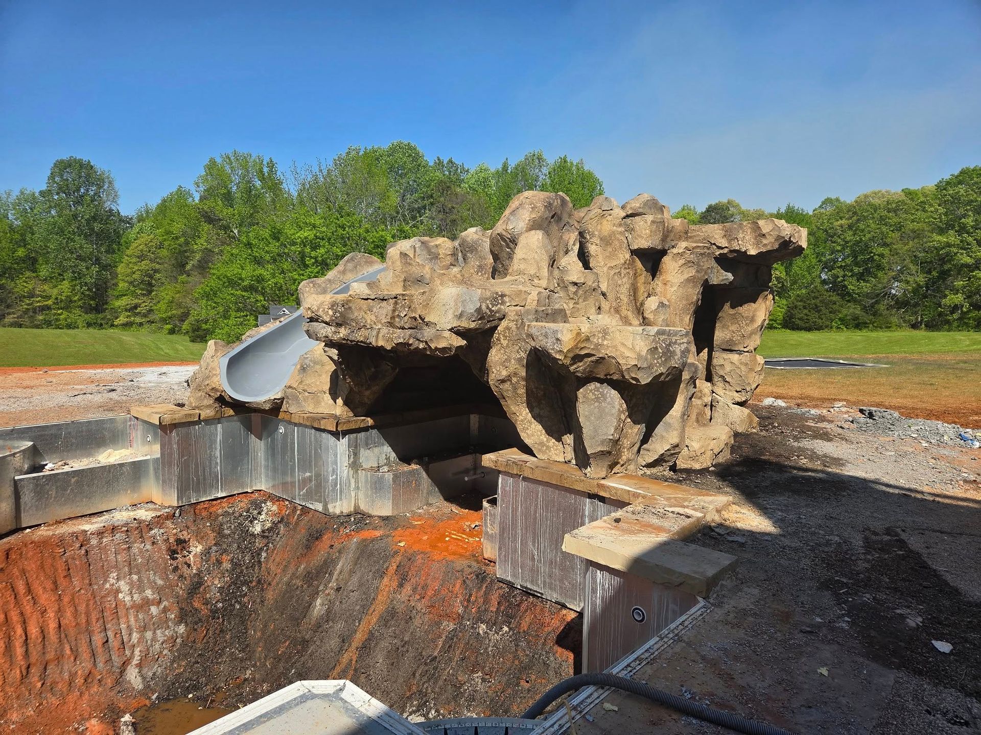 A faux rock water feature with a slide, perched above a pool under construction, with a forest background.