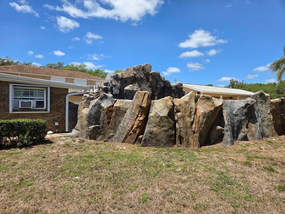 Large rock formation in front of a house, set in a yard with grass and blue sky.