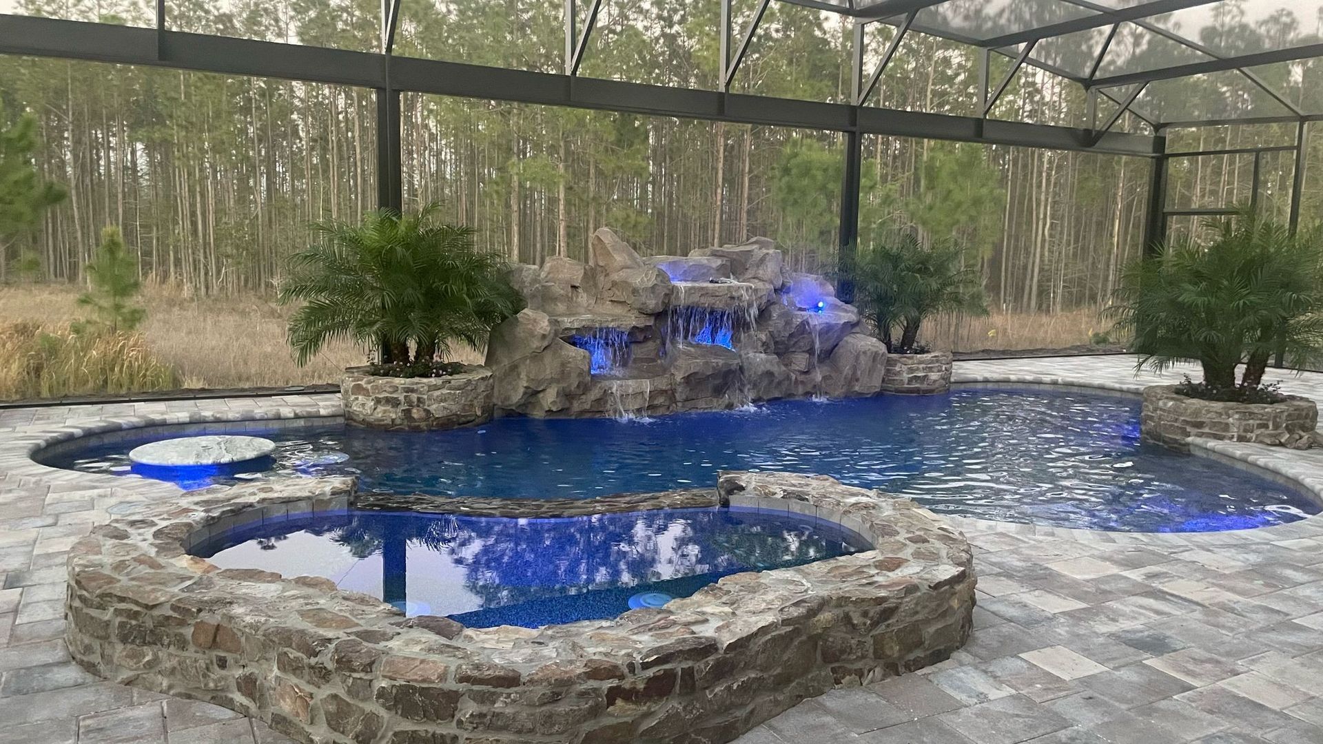 Swimming pool with waterfall feature, surrounded by stone and lush landscaping under a screened enclosure.