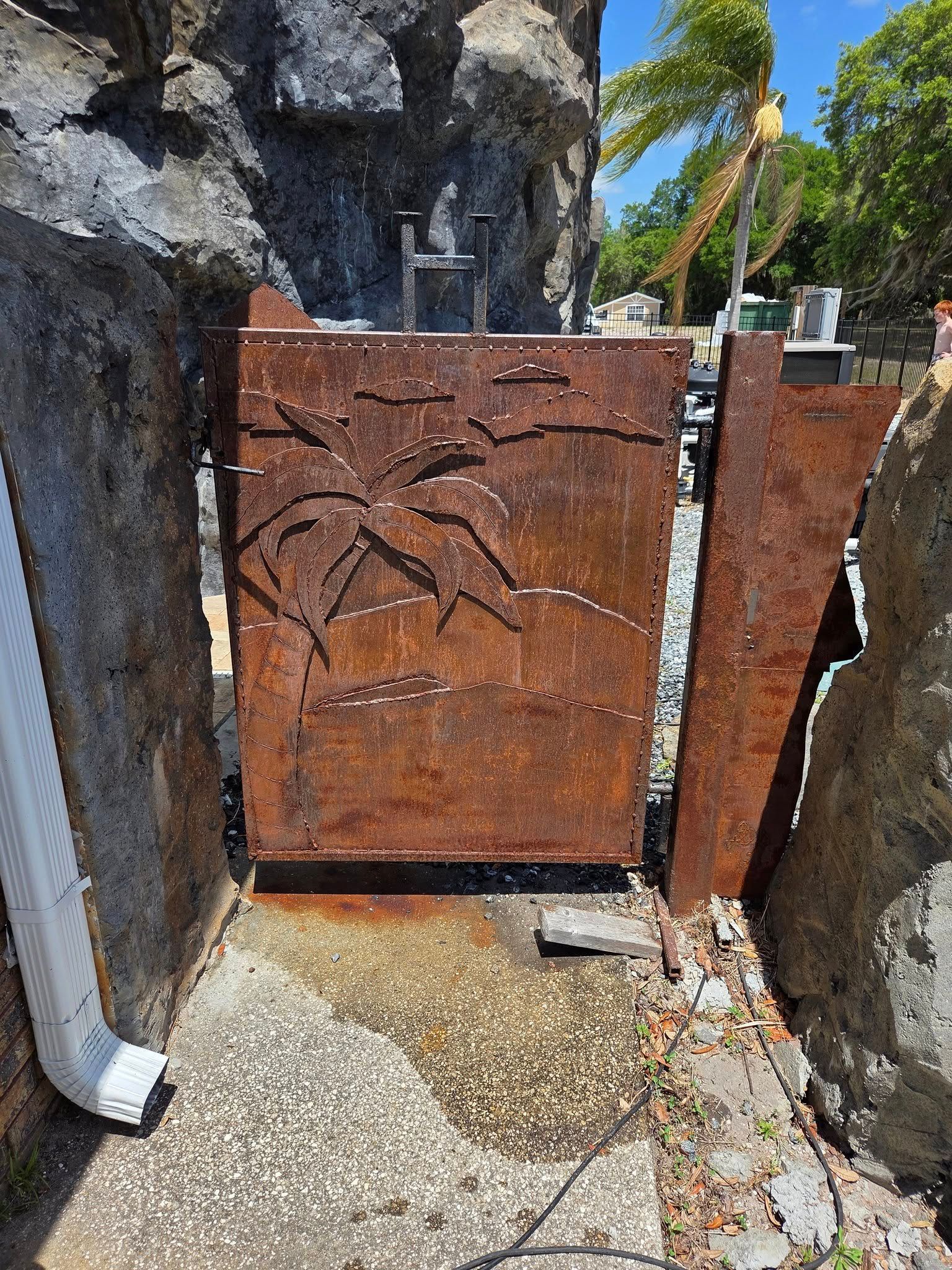 Rusty metal gate with palm tree design, between rock walls. Water stains the pavement.