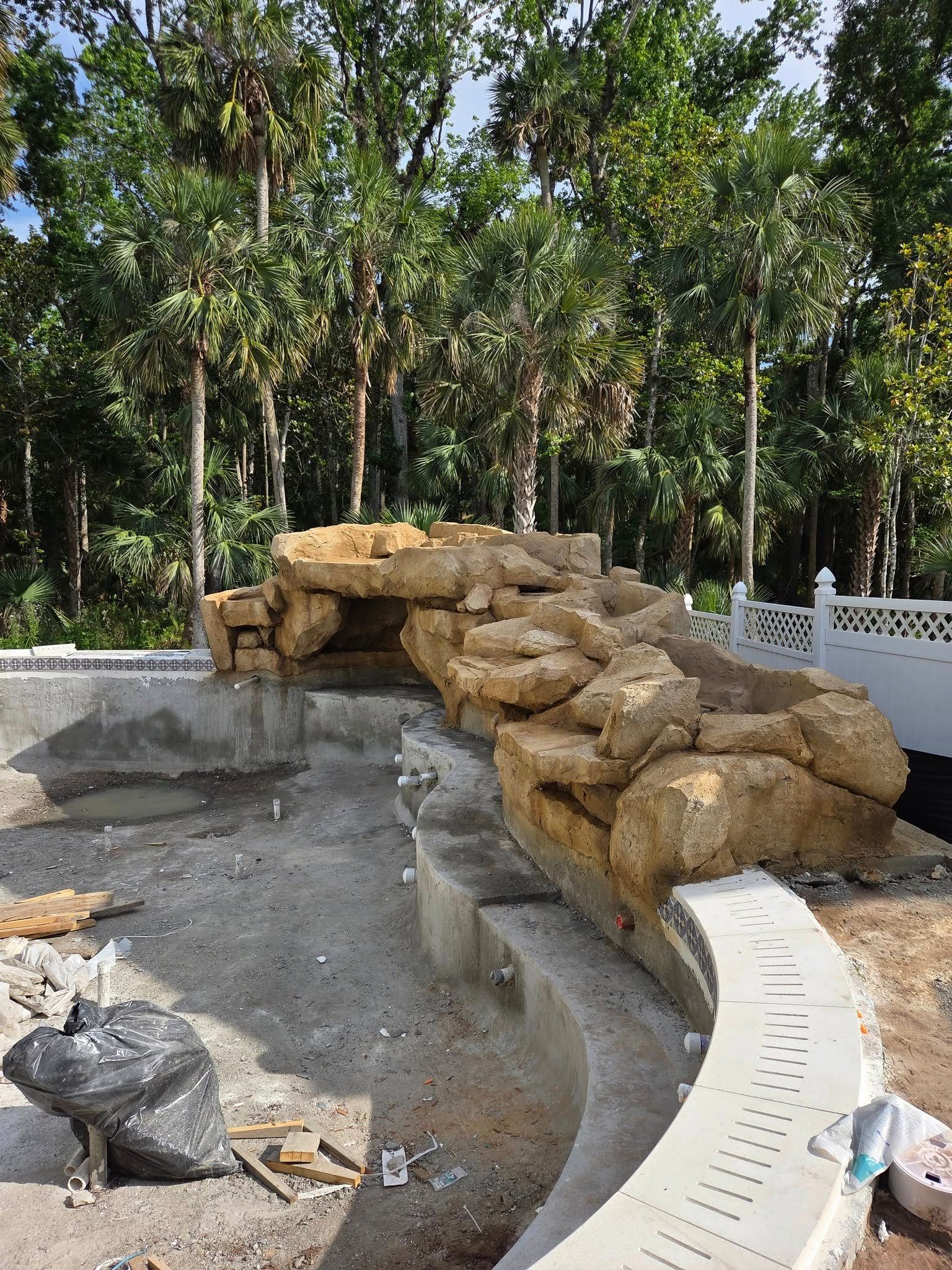 Pool construction with rock waterfall feature, concrete, trees.