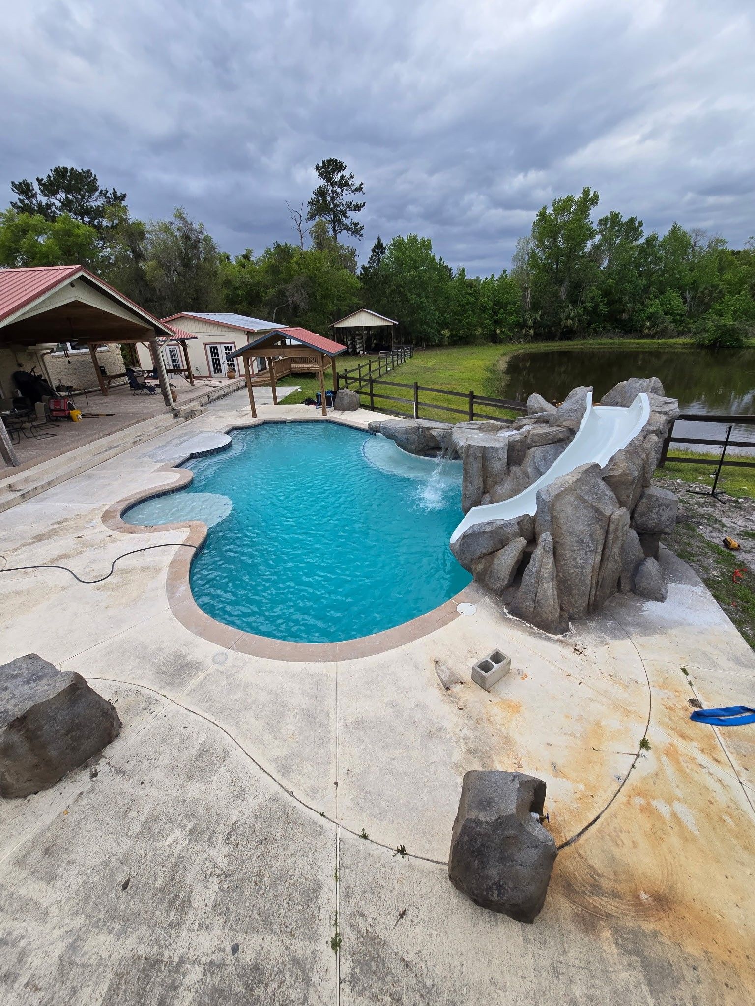 Pool with rock slide, patio, and lake under a cloudy sky.