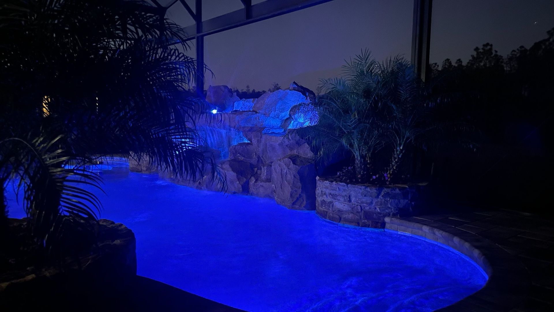 Blue illuminated swimming pool at night with a rock waterfall feature and surrounding plants.