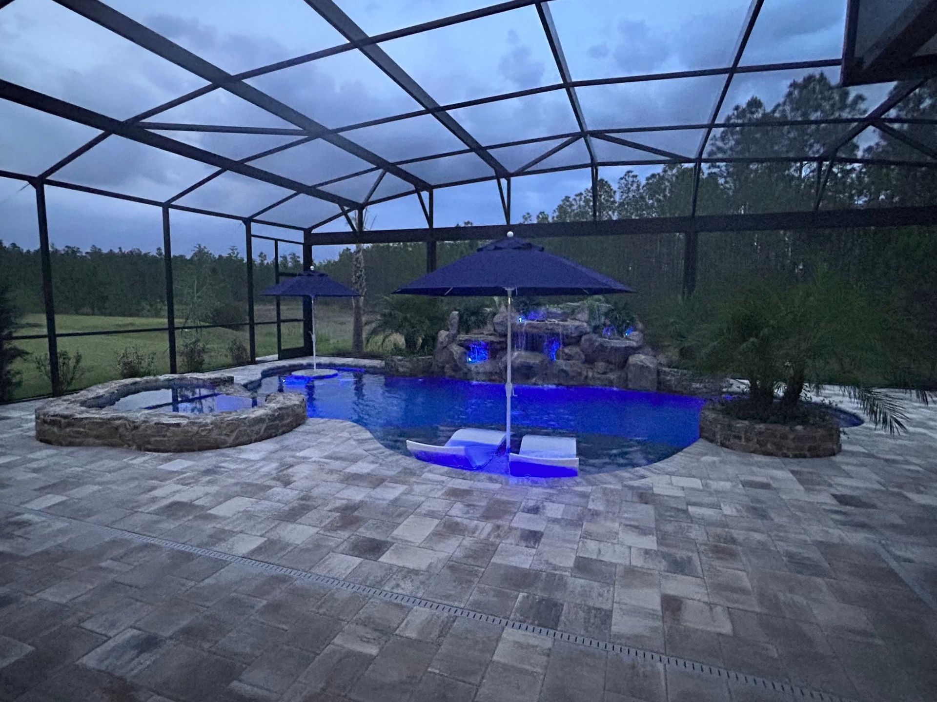 Pool under a screened enclosure with blue lights, umbrellas, and stone patio.