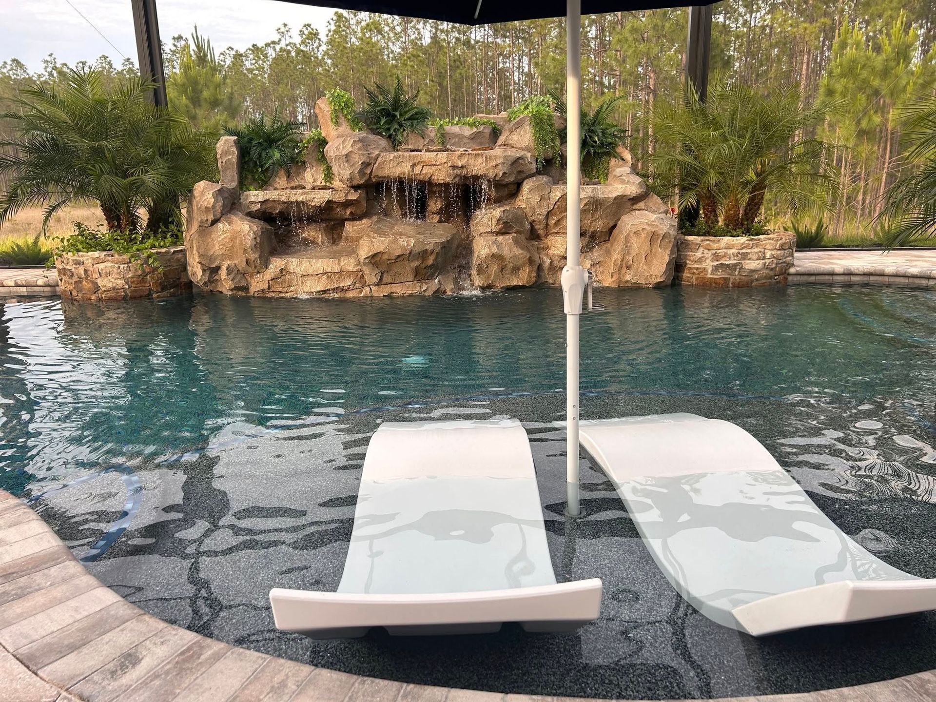 Pool with waterfall, two in-pool chaise lounges, and black umbrella on a sunny day.