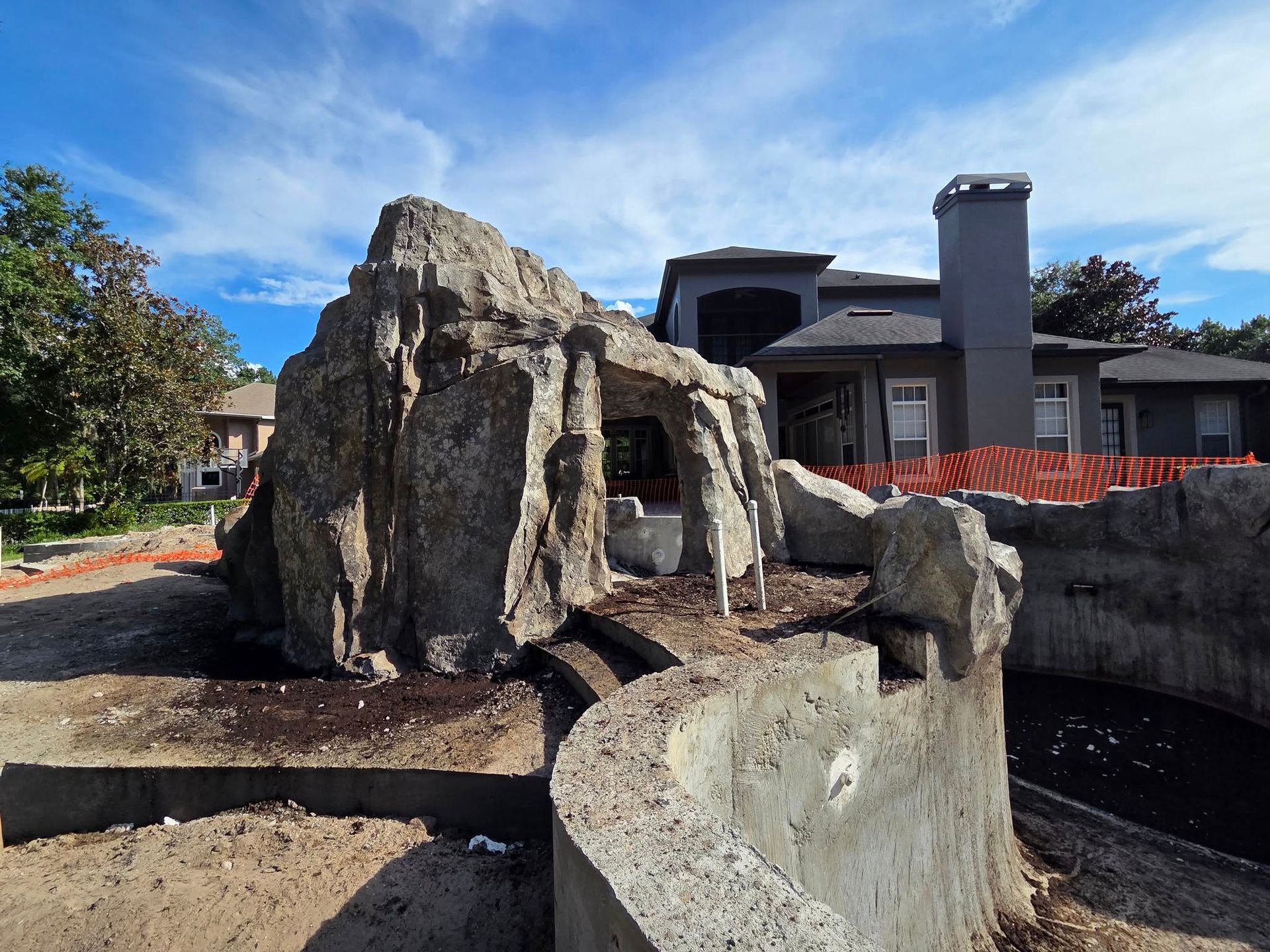 A concrete rock formation with a cave-like opening sits next to a pool under construction, with a house in the background.