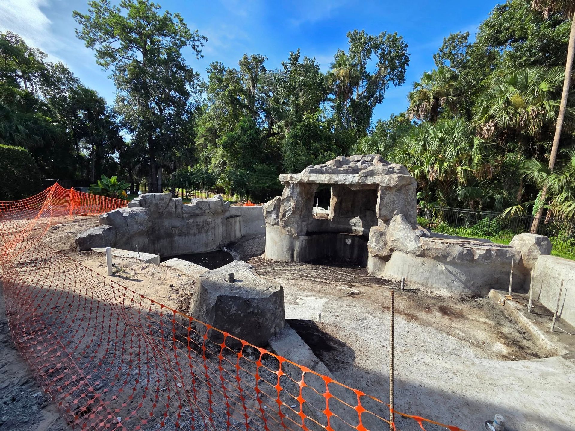 Ruined concrete structure under construction, surrounded by orange fencing and trees on a sunny day.