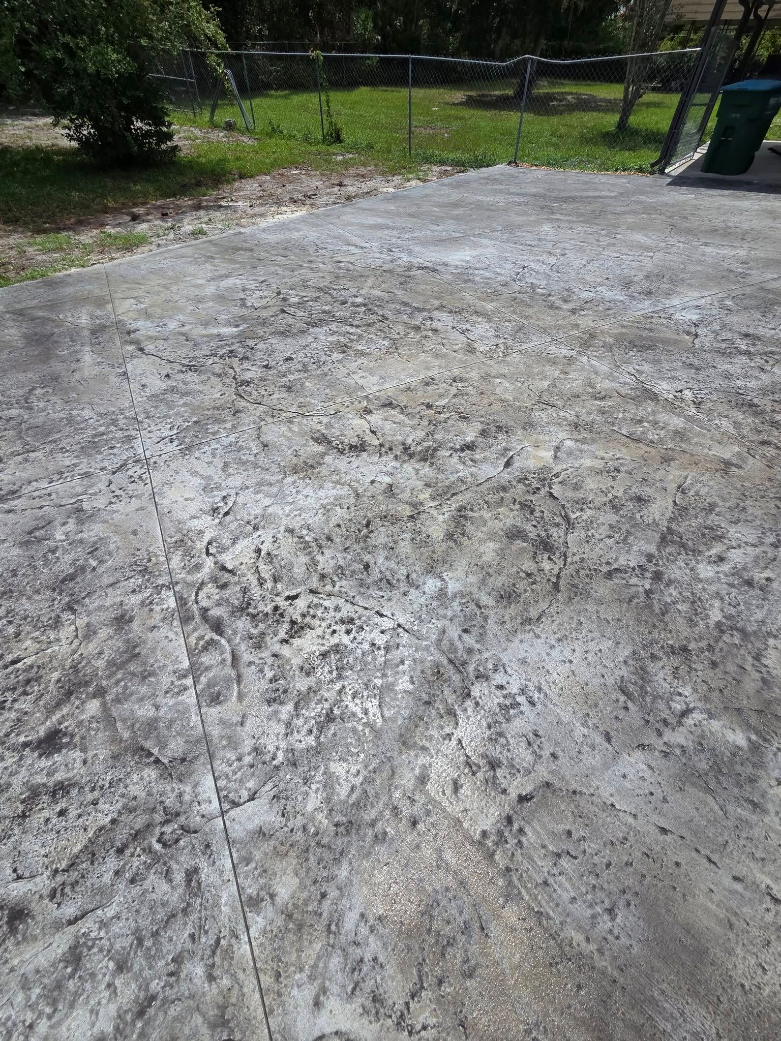 Weathered concrete patio with discoloration, cracks, and some debris. Green grass and fence in the background.
