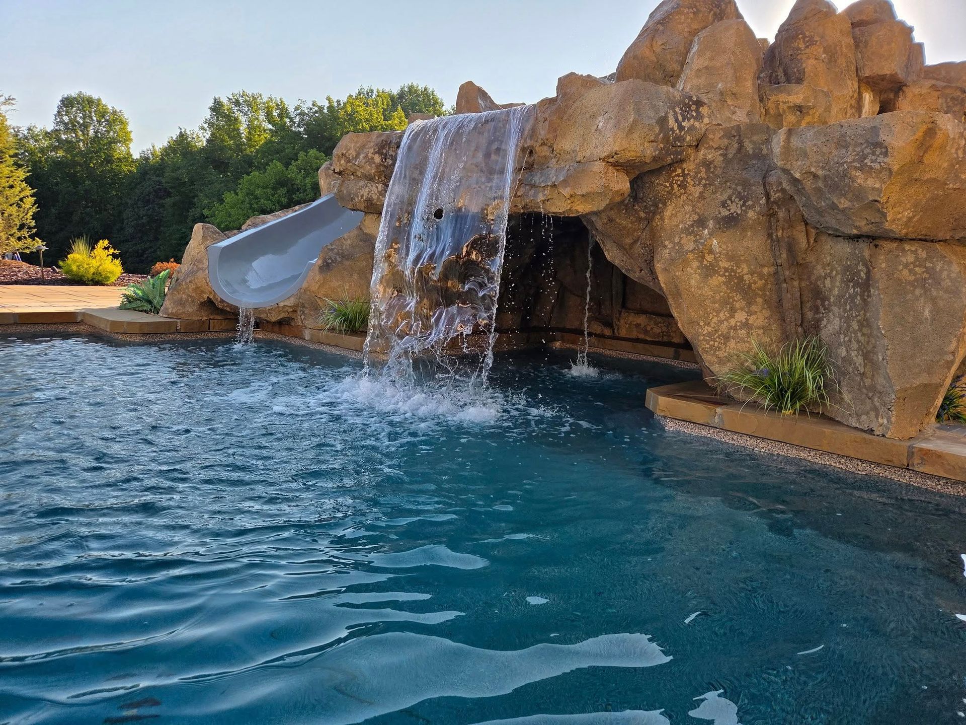 Pool with waterfall feature and slide, surrounded by rock structure, trees in the background, blue water.
