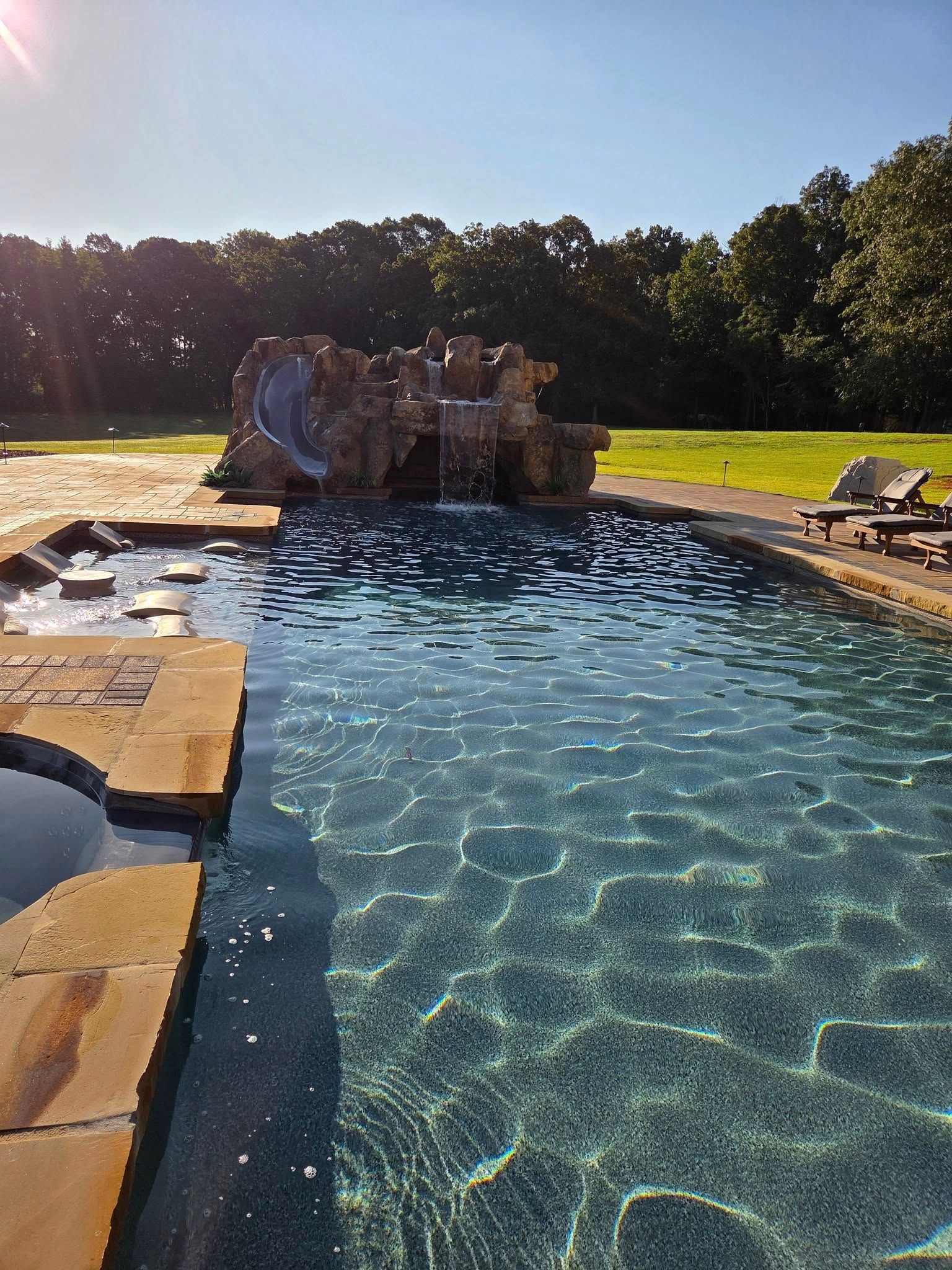 Swimming pool with rock waterfall and slide, sunny outdoor setting.