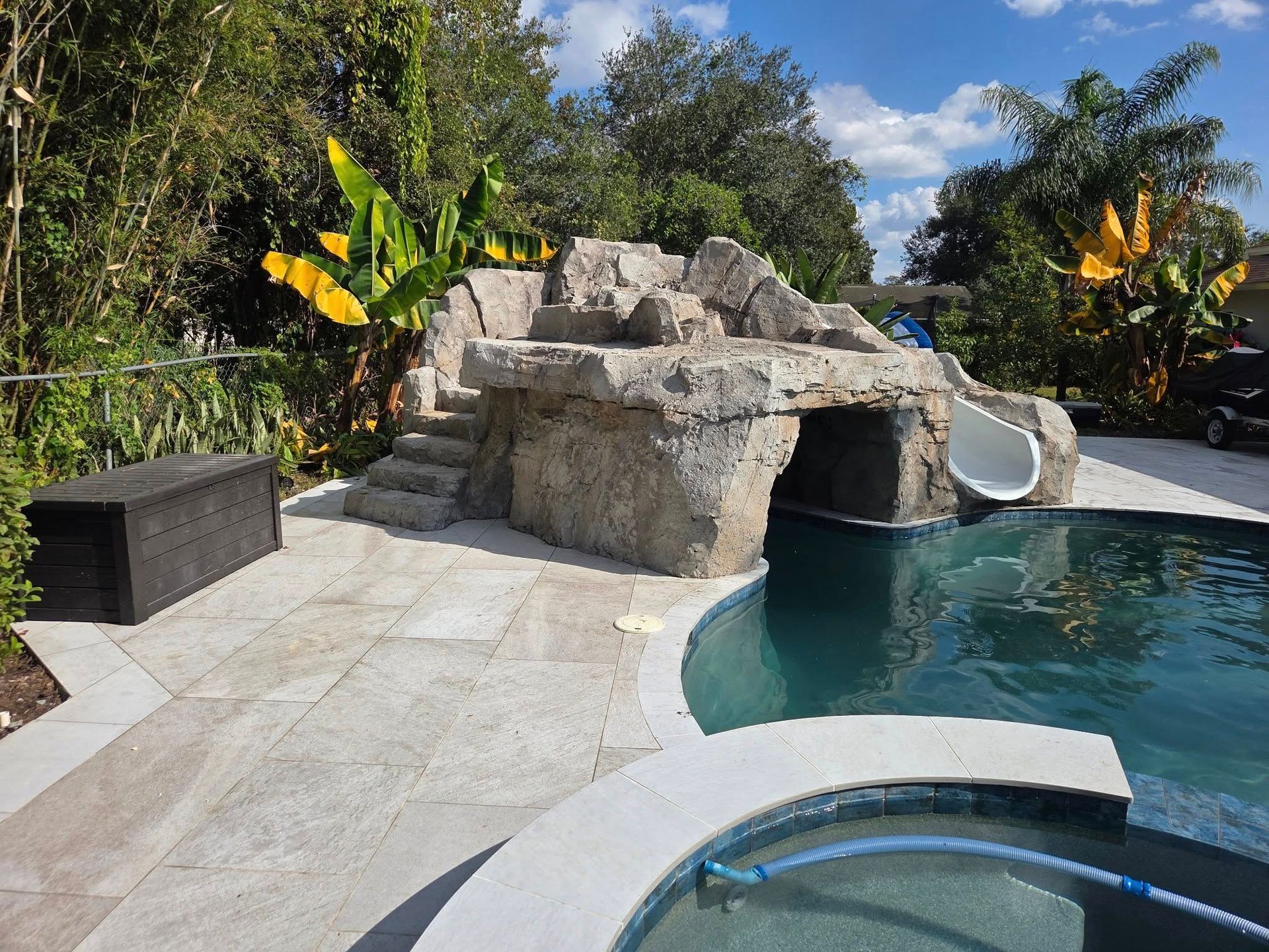 Backyard pool with rock feature, slide, steps, and spa. Blue water, stone patio, green foliage, and sunny sky.