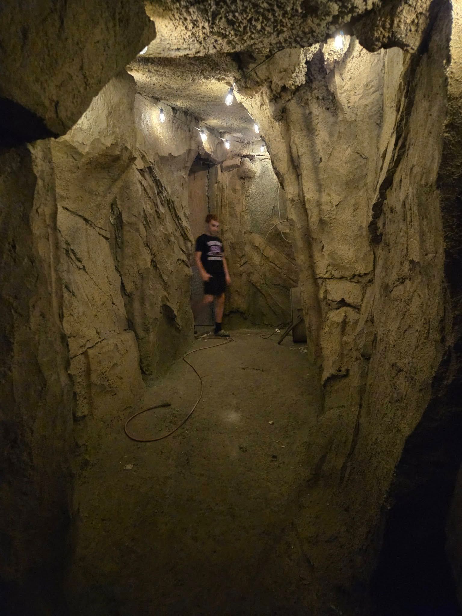 Person stands in a dimly lit, narrow tunnel with rough, textured walls. Overhead lights illuminate the space.