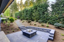 Stone patio with fire pit, steps, and pathway leading to landscaped yard with evergreen trees.