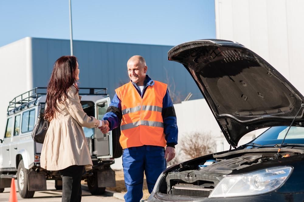 A Man In An Orange Vest Is Shaking Hands With A Woman In Front Of A Broken Down Car — Affordable Mechanic in Albury, NSW