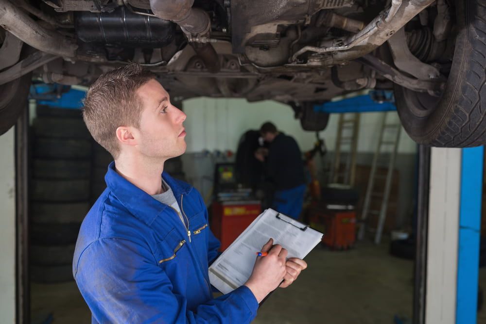 A Man Is Looking Under A Car In A Garage While Holding A Clipboard — Vehicle Inspections & Mechanical Services in Albury, NSW