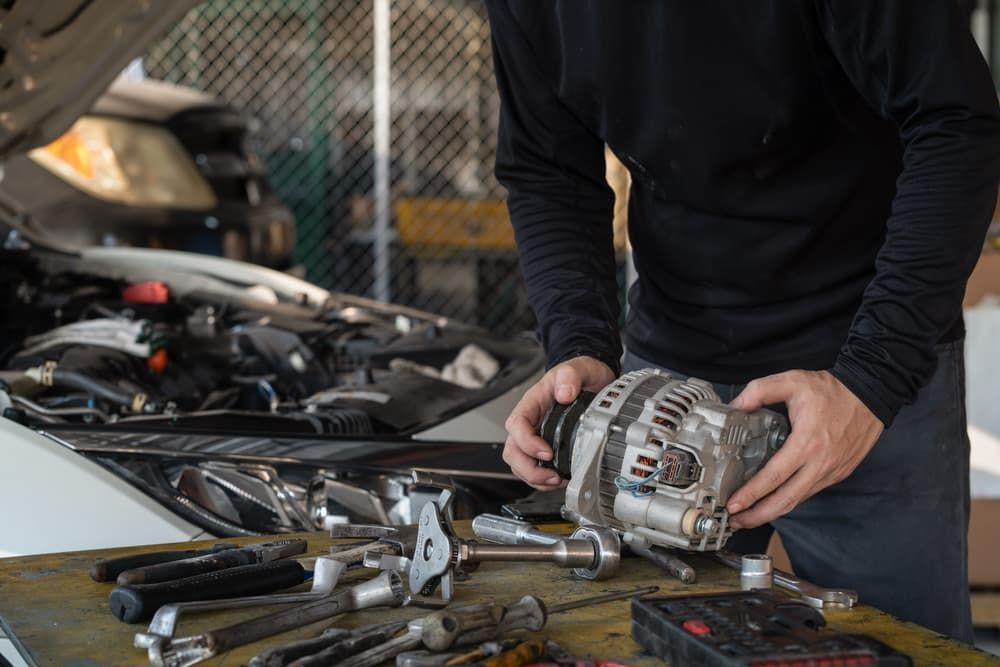 A Man Is Holding A Car Alternator In Front Of A Car With The Hood Open — Experienced Car Mechanic in Albury, NSW