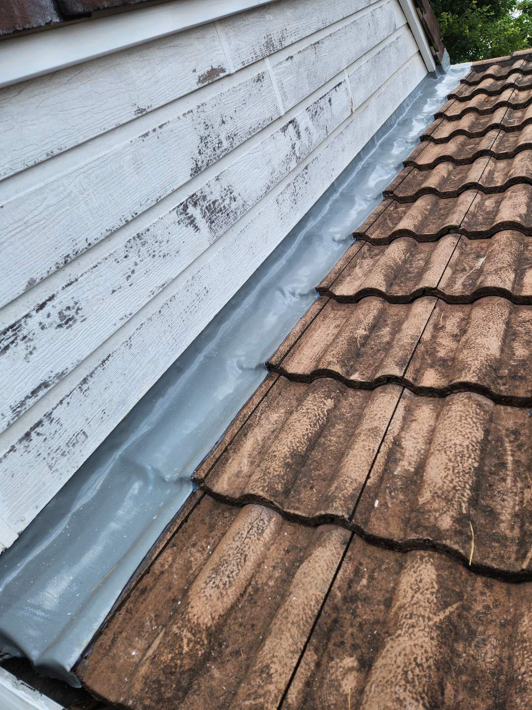 A section of a roof where weathered white siding meets brown clay tiles, sealed with a gray metal flashing strip.